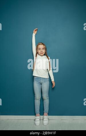 Little blonde girl smiling and measuring height against blue wall in room Stock Photo