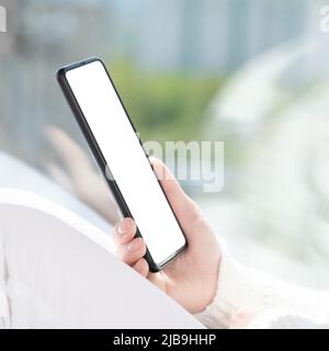 Cell phone mockup. Girl in white clothes sits by window and uses cell phone, looks at smartphone, reads, shop, communicates with friends in messenger. Stock Photo
