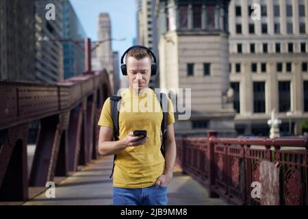 Young man listening to music with wireless headphones and looking into his cell phone during city walk. Stock Photo