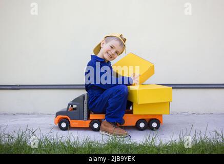 cute smiling 6 year old boy in uniform sits on a big toy car - a truck with many cardboard boxes. Parcel delivery, little postman, truck driver. Posit Stock Photo