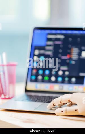 Robotic arm using laptop on a wooden table. Artificial intelligence robot working on an open laptop. Futuristic robot hand typing and working with lap Stock Photo