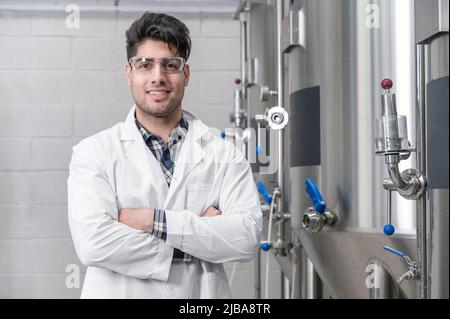 Young Handsome male worker in white lab coat smiling and looking to camera, standing at rows of steel brewing vats and crossing hands .Concept of brewery and beer plant. High quality photography. Stock Photo