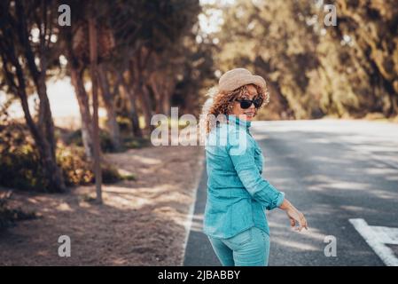 Happy woman viewed from back looking on camera and smile walking on the road with trees on both side. Tourist with denim casual clotehs and straw hat Stock Photo