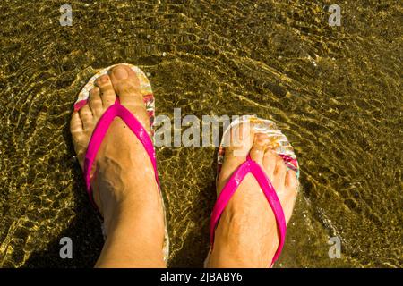 Feet of an unknown woman, wearing flip flops, on the beach in the sea Stock Photo