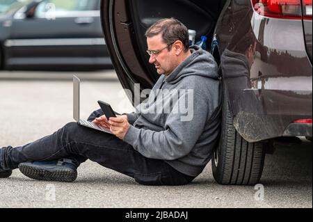 middle-aged man sitting on the asphalt by the car and working on a laptop, working remotely, travel Stock Photo