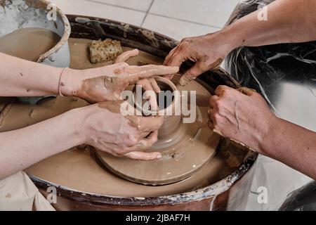 Hands of senior woman and girl sculpting clay vase on potter's wheel at pottery training lesson. Stock Photo