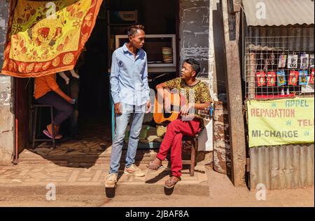 Antananarivo, Madagascar - May 07, 2019: Two unknown Malagasy men chatting in front of small shop at main street, sitting one holding guitar Stock Photo