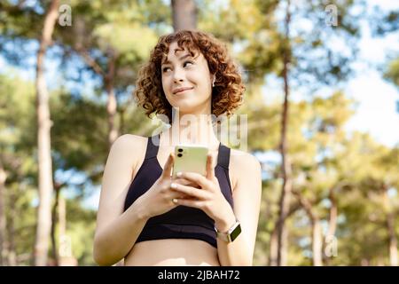 Portrait of young redhead woman wearing sportive clothes on city park, outdoors holding and using smart mobile phone and lookin away. Thinking dreamin Stock Photo