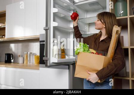 Woman with a paper shopping bag full of vegetables in the kitchen puts groceries in the fridge Stock Photo