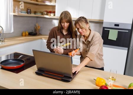 Middle aged woman and her adult daughter cooking in front of a laptop in the kitchen Stock Photo