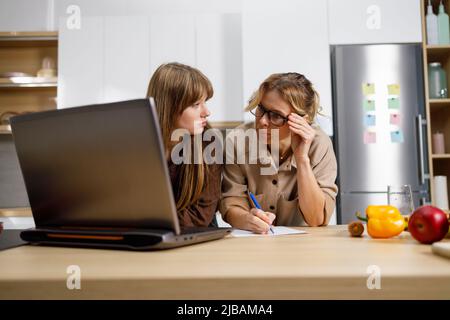 Adult woman and her daughter in the kitchen writing something while sitting in front of a laptop Stock Photo
