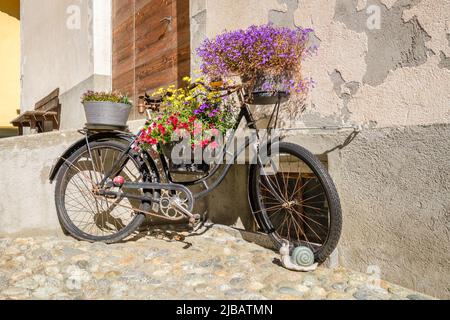 An old rusty bike, decorated with beautiful flowers, in the historic village center of S-Chanf (Upper Engadine Valley , Grisons, Switzerland) Stock Photo