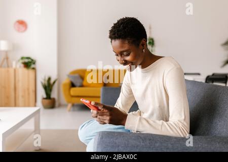 Happy young african woman using cellphone sitting on sofa at home Stock Photo