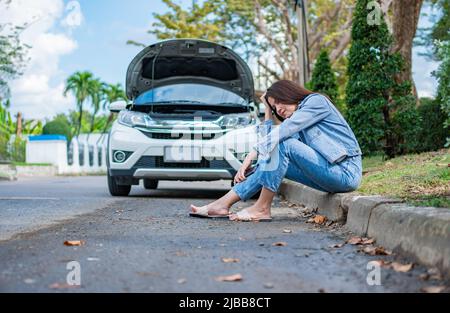 Asian woman sitting beside car after a car breakdown on street. Concept of vehicle engine problem or accident and emergency help from Professional mec Stock Photo