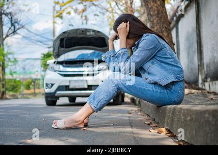 Asian woman sitting beside car after a car breakdown on street. Concept of vehicle engine problem or accident and emergency help from Professional mec Stock Photo