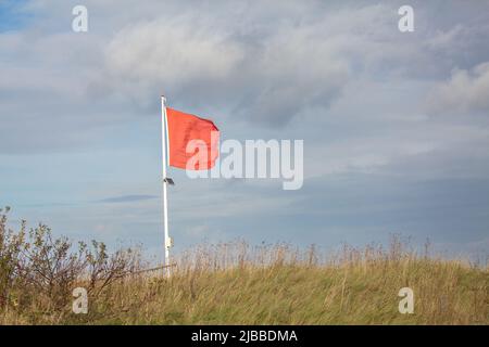 Red flag waving in the wind with small solar panel on the post at Norfolk grass beach in UK Stock Photo