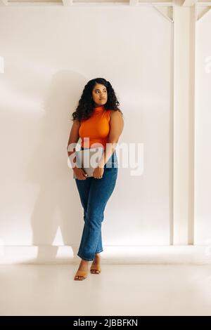 Young businesswoman looking away thoughtfully while standing against a wall background. Pensive female entrepreneur holding a laptop in a modern workp Stock Photo