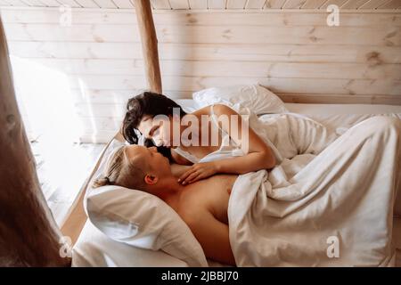 A young man and woman are basking in bed together. They are lying under a white sheets. A loving couple just woke up. Look at each other with tenderness. Stock Photo