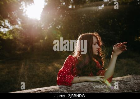 Portrait of pretty redhead woman in red summer loose shirt leaning by hands on a dry beam in wood and posing on a camera Stock Photo