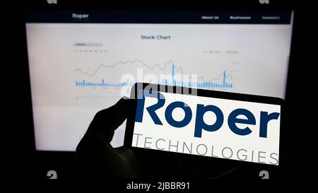 Person holding mobile phone with logo of US engineering company Roper Technologies Inc. on screen in front of web page. Focus on phone display. Stock Photo