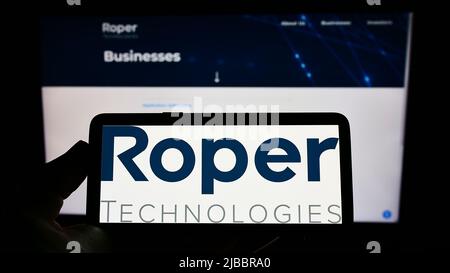 Person holding cellphone with logo of American engineering company Roper Technologies Inc. on screen in front of webpage. Focus on phone display. Stock Photo