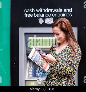 Epsom Surrey, London UK, June 04 2022, Woman Standing Reading Newspaper, ATM Cashpoint Machine In Background Stock Photo