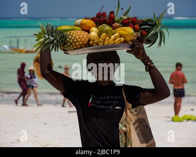 Paje, Zanzibar, Tanzania - January 202: African man with a tray full of exotic fruits on his head. Turquoise ocean water in the background. Africa Stock Photo