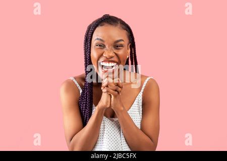 Emotional young black woman in summer dress clasping hands together in excitement on pink studio background Stock Photo