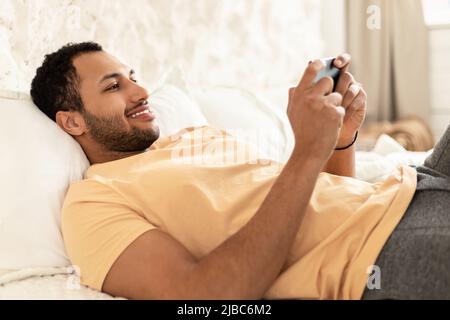 Cheerful Arabic Guy Using Cellphone Playing Game Lying At Home Stock Photo