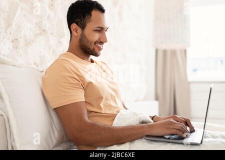 Happy Arabic Male Freelancer Using Laptop In Bedroom, Side View Stock Photo