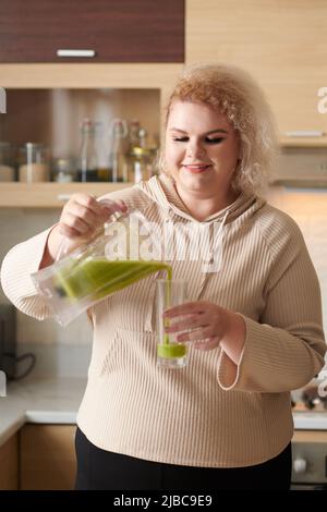 Portrait of happy smiling young woman pouring green smoothie she made for breakfast in glass Stock Photo
