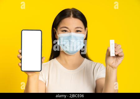 Happy asian woman wearing medical face mask, holding smartphone and test, screen showing app interface, health, vaccination passport, certificate. Call your doctor concept. Product placement mock up.  Stock Photo