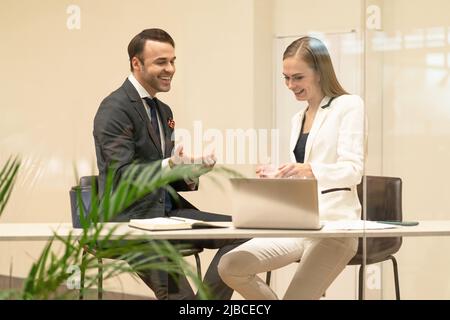 Two business partners discuss updates while having a coffee break. Young business people in the suits with coffee in hands looking down standing behind the glass.  Stock Photo