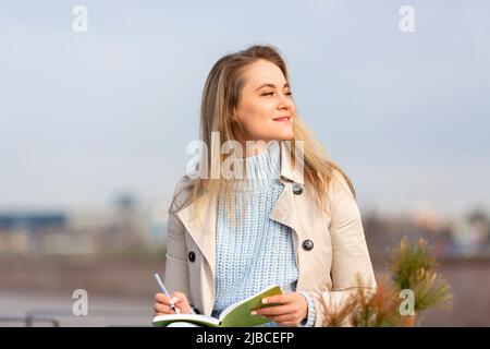 A young woman makes notes in a notebook during a warm spring day. She smiles and looks away. Stock Photo