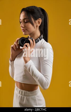 Thoughtful woman in casual clothes holding headphones hanging on neck. Side view of cute girl with ponytail wearing earphones, while looking down, isolated on orange background. Concept of lifestyle. Stock Photo
