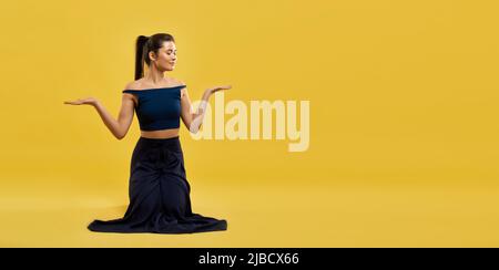 Smiling woman standing on knees keeping hands palms up indoor. Front view of graceful girl in black wear kneeling with arms bent at elbows, isolated on yellow studio background. Concept of posture. Stock Photo