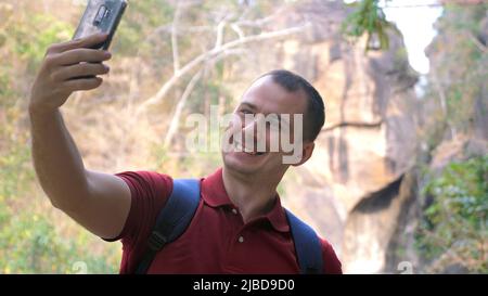 Smiling male traveler takes selfies on phone against canyon in a national park. Happy male hiker records video or takes photos with front-facing Stock Photo