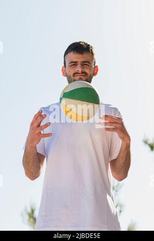 A young Caucasian man with a beard is passing a small, multicolored basketball from one hand to the other. The photograph is taken in backlight. Stock Photo