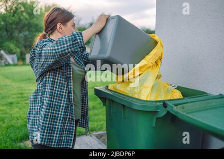 European 30s woman throwing garbage into the recycling bin in the backyard near the house. Separating recyclable garbage plastic  Stock Photo