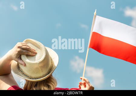 A young woman proudly holds the Polish flag above her head, against the sky with her other hand holding her hat, Concept of national holidays and patr Stock Photo