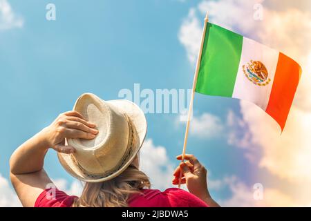 A young woman proudly raises the flag of Mexico above her head against the sky, The other hand holds up her hat, Concept for national holidays and pat Stock Photo
