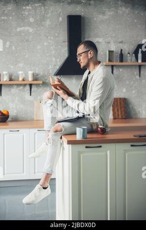 Young man using tablet remote working from home, making notes in notebook sitting in the kitchen. Contemporary portrait. Remote workplace. Stock Photo