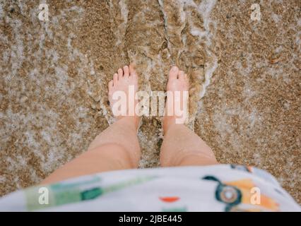 womans feet and legs stood in the sand with the sea splashing Stock Photo