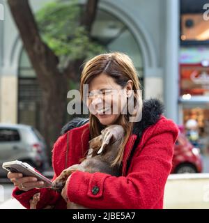 Latin woman using her smartphone and laughing while holding her dog Stock Photo