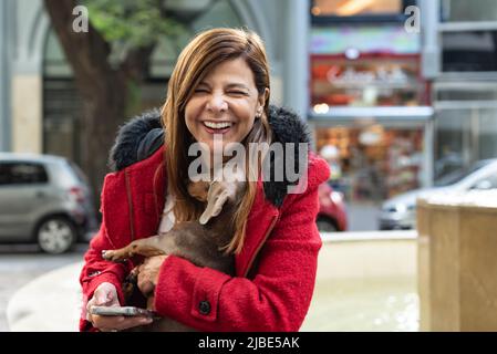 Latin woman laughing while holding her dog and smartphone Stock Photo