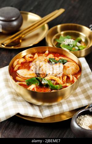 Red Seafood Jjampong, Red Spicy Seafood Korean Noodle Soup with Vegetable and Clam on Golden Bowl, Above Wooden Table Stock Photo