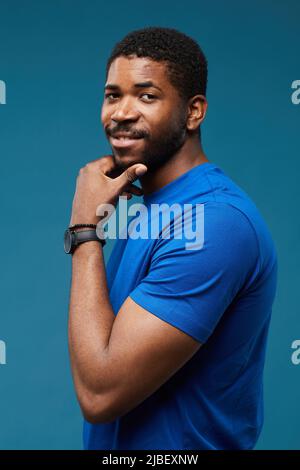 Vertical portrait of handsome black man looking at camera over shoulder with smug expression and hand on chin while wearing blue on blue background Stock Photo