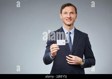Waist up portrait of smiling businessman showing credit card mockup to camera against grey background, copy space Stock Photo