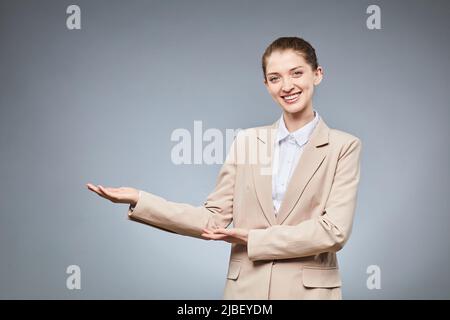 Waist up portrait of smiling young businesswoman pointing at copy space while standing against grey background Stock Photo