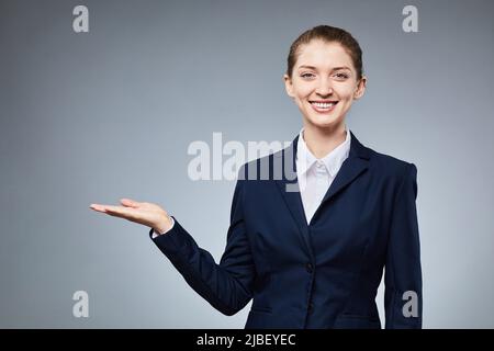 Waist up portrait of young businesswoman pointing at copy space and smiling at camera while standing against grey background Stock Photo